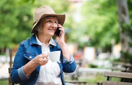 Senior woman with hat sitting outdoors in cafe, using smartphone.