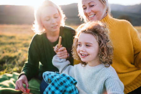 Small girl with mother and grandmother having picnic in nature at sunset.
