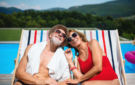 Cheerful senior couple sitting by swimming pool outdoors in backyard.の写真素材