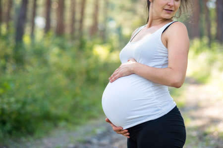 Unrecognizable pregnant woman outdoors in nature, touching her belly.