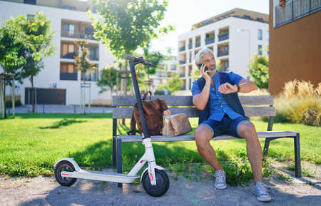 Portrait of mature man with electric scooter sitting outdoors in city, using smartphone.の写真素材