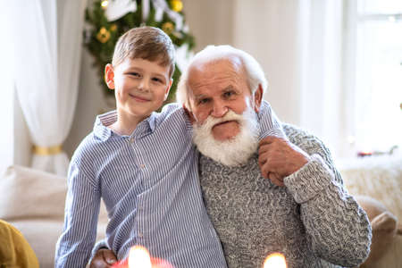 Small boy with senior grandfather indoors at home at Christmas, looking at camera.