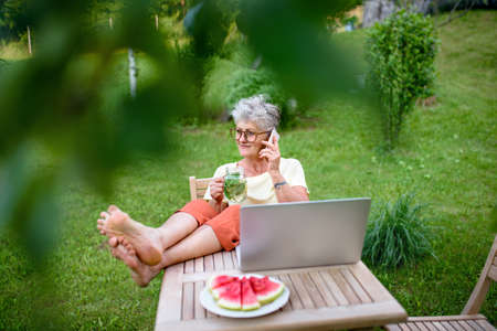 Happy senior woman with laptop and smartphone working outdoors in garden, home office concept.