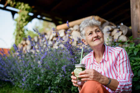 Portrait of senior woman sitting outdoors in garden, holding lemonade drink.