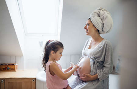 Small girl creaming belly of pregnant mother indoors in bathroom at home.