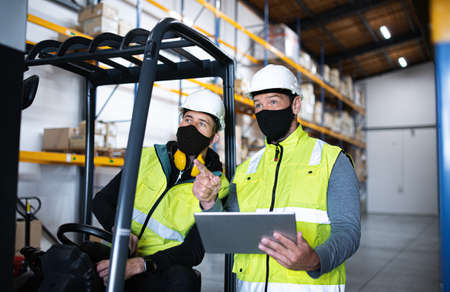 Workers with face mask and tablet working indoors in warehouse, coronavirus concept.の写真素材