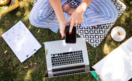 Unrecognizable woman working in home office outdoors in garden, using smartphone and laptop.の写真素材
