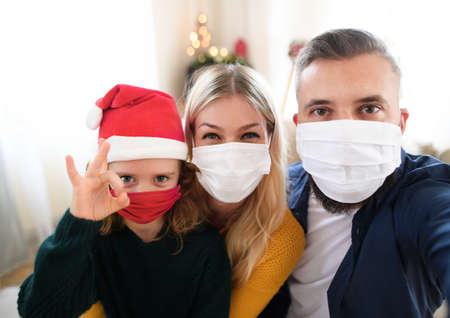 Family with small daughter indoors at home at Christmas, taking selfie with face masks.