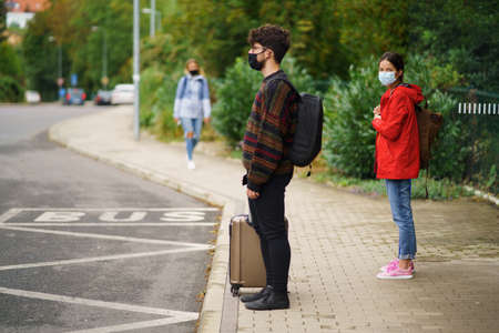 Young people with bags waiting on bus stop outdoors in town. Coronavirus concept.