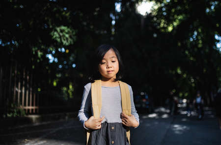 Portrait of small Japanese girl with backpack standing outdoors in town.