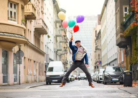 Happy senior man with balloons outdoors on street in city, jumping.