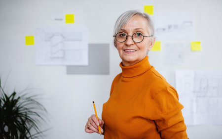 Portrait of senior woman architect standing indoors at home, working.