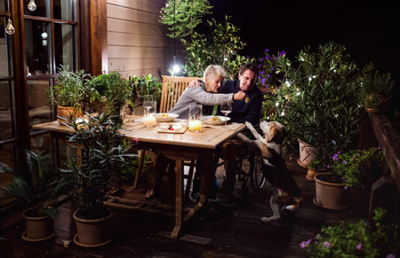 Senior couple in wheelchair having dinner in the evening on terrace, feeding dog.の写真素材