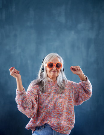 Senior woman with sunglasses and headphones standing indoors against dark background, dancing.の写真素材