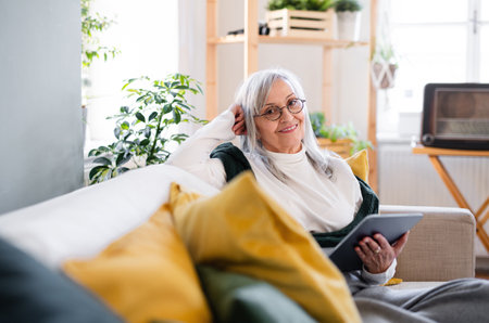 Portrait of senior woman sitting indoors on sofa at home, using tablet.