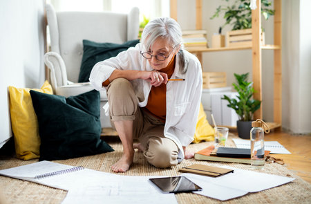Portrait of senior woman architect sitting on floor indoors at home, working.