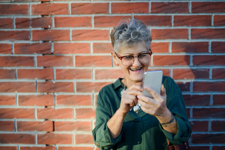 Senior woman standing outdoors against brick wall background, using smartphone.