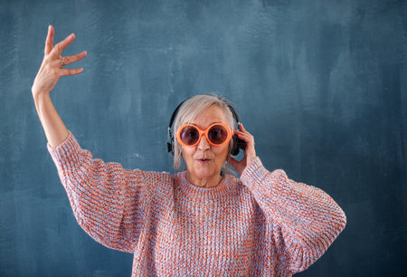 Senior woman with sunglasses and headphones standing indoors against dark background.の写真素材