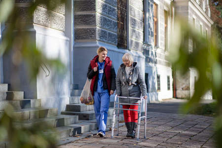 Senior woman with walking frame and caregiver outdoors in town, shopping and coronavirus concept.