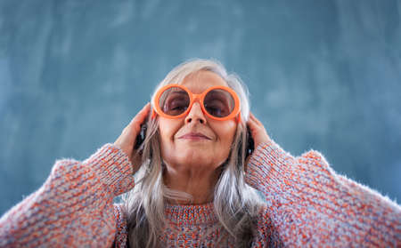 Portrait of senior woman with sunglasses standing indoors against dark background, listening to music.の写真素材