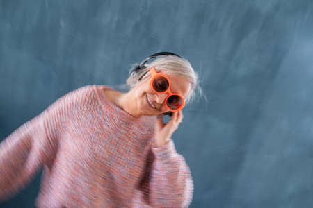 Senior woman with sunglasses and headphones standing indoors against dark background.