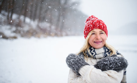 Front view portrait of senior woman with hat and mittens outdoors standing in snowy nature.