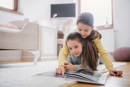 Two small girls sisters indoors in bedroom at home, reading a book.の写真素材