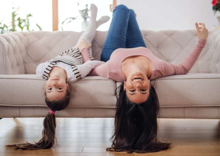 Mother with small daughter on sofa indoors at home, looking at camera upside down.の写真素材