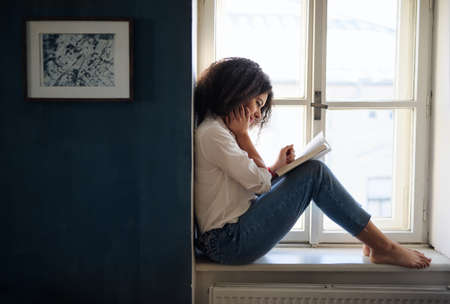Portrait of young woman with book on window sill indoors at home, reading.