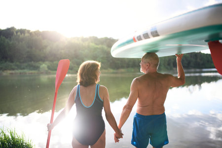 Rear view of senior couple carrying paddleboard by lake in summer.の写真素材