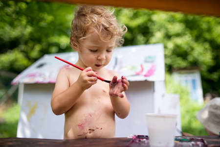 Topless small blond girl painting paper house outdoors in summer.の写真素材