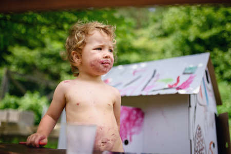 Topless small blond girl painting paper house outdoors in summer.の写真素材