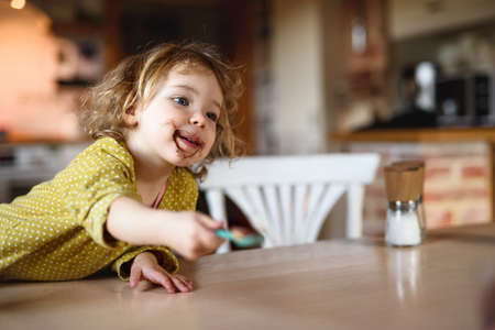 Small girl with dirty mouth indoors in kitchen at home, eating pudding.の写真素材
