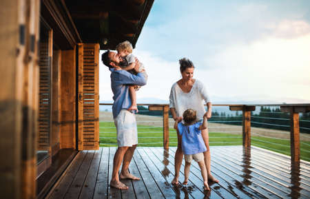 Family with small children playing in rain on patio by wooden cabin, holiday in nature concept.の写真素材