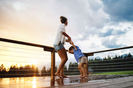 Mother with small daughter playing in rain on patio of wooden cabin, holiday in nature concept.の写真素材