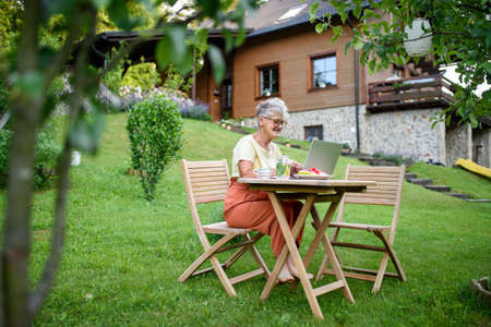 Happy senior woman with laptop working outdoors in garden, home office concept.の写真素材