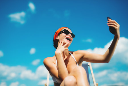 Young woman sitting by swimming pool outdoors in backyard garden, taking selfie.の写真素材