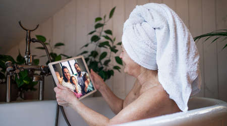 Senior woman with tablet in bath tub at home, video call with daughter and grandchildren.