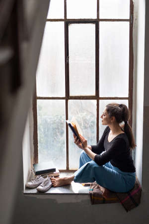 Happy woman sitting on old and dirty window sill, reading a book.の写真素材