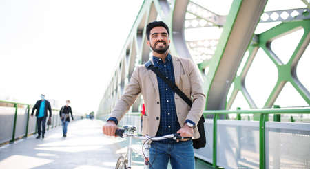 Young business man commuter with bicycle going to work outdoors in city, walking on bridge.の写真素材