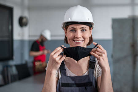 Portrait of woman worker with helmet indoors in factory putting on face mask.の写真素材