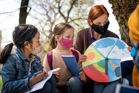 Teacher with small children sitting outdoors in city park, learning group education and coronavirus...