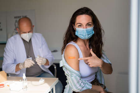 Happy young woman after covid-9 vaccination, pointing to plaster on arm and looking at camera.