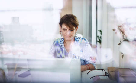 Mature woman with laptop working indoors in home office, small business concept. Shot through glass.の写真素材