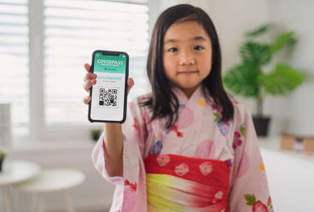 Small japanese child with traditional costume showing covid-19 vaccination certificate on...