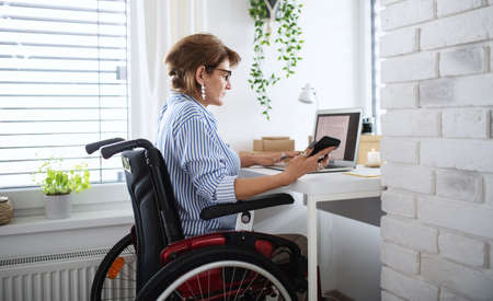 Disabled mature woman with laptop working indoors in home office, small business concept.