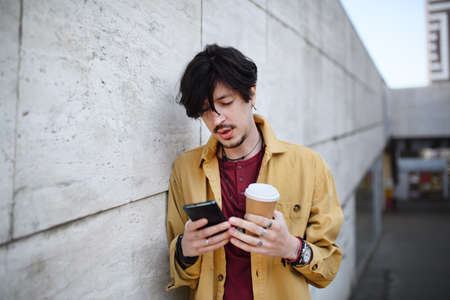 Portrait of young man standing against concrete wall outdoors, using smartphone.の写真素材