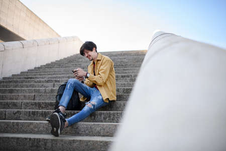 Portrait of young man sitting on staircase outdoors in city, using smartphone.