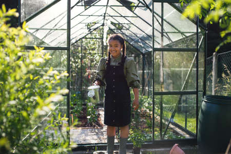 Happy small girl gardening in greenhouse outdoors in backyard, looking at camera.