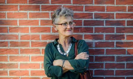 Senior woman standing outdoors against brick wall in town, arms crossed.